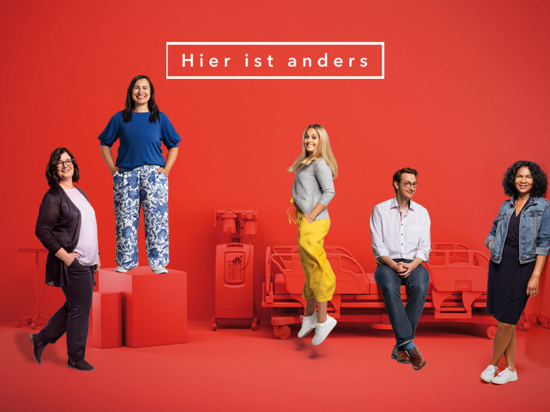 Bild mit der Aufschrift „Hier ist anders.“. Vier Frauen und ein Mann in gelassener Atmosphäre vor einem Patientenbett. Der Hintergrund ist rot. 