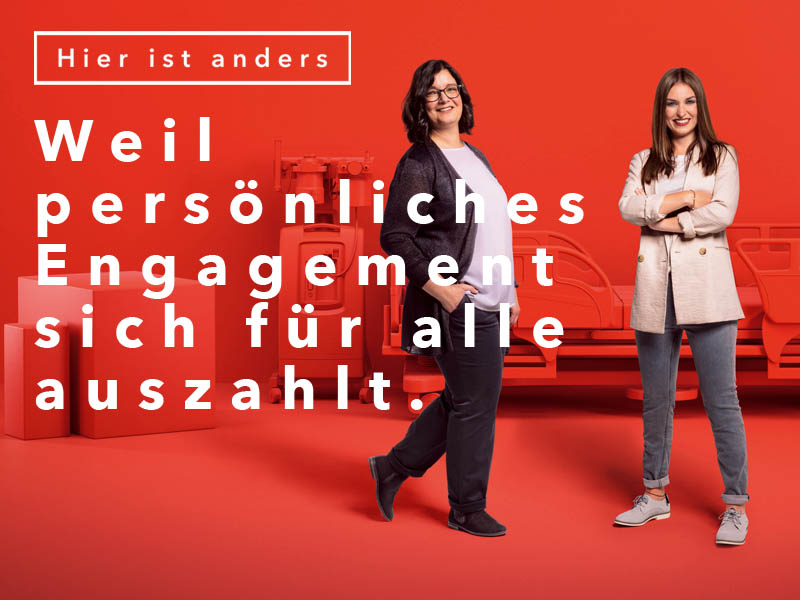 Bild mit der Aufschrift „Weil persönliches Engagement sich für alle auszahlt.“. Zwei lächelnde Frauen stehen vor einem Patientenbett. Der Hintergrund ist rot. 